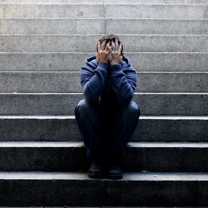 Young desperate man in casual clothes abandoned lost in depression sitting on ground street concrete stairs alone suffering emotional pain, sadness, looking sick in grunge lighting